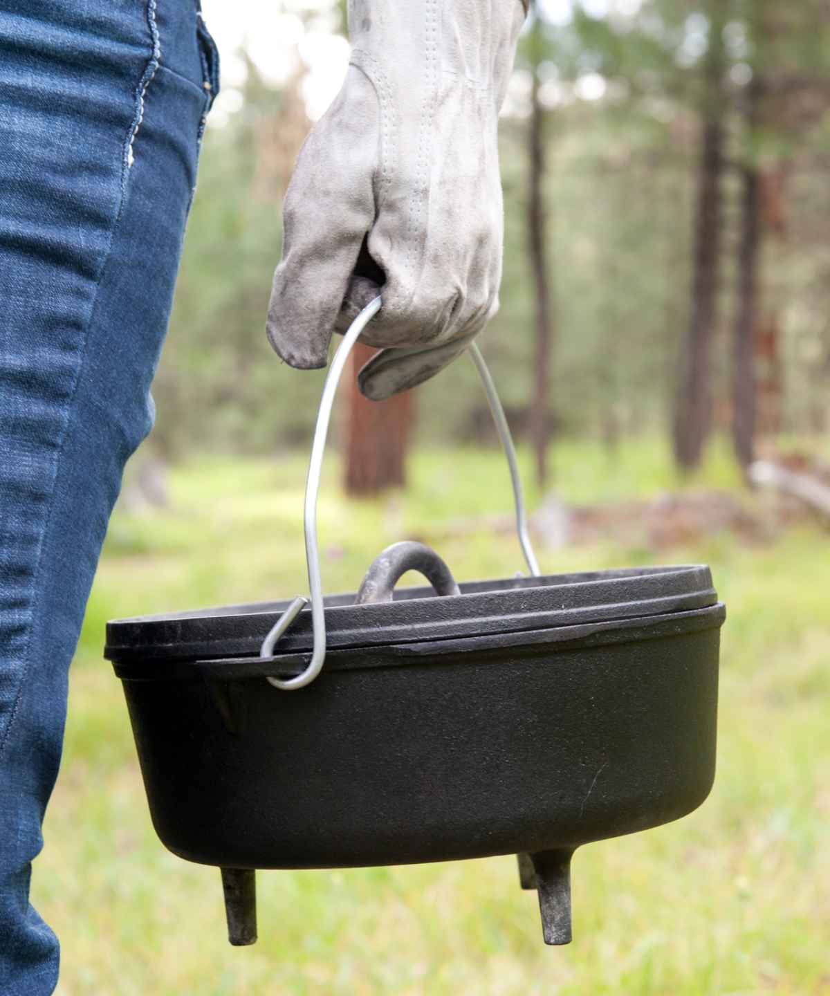 Nahaufnahme eines Dutch Ovens, der von einer Person mit Handschuh getragen wird, im Hintergrund Bäume.