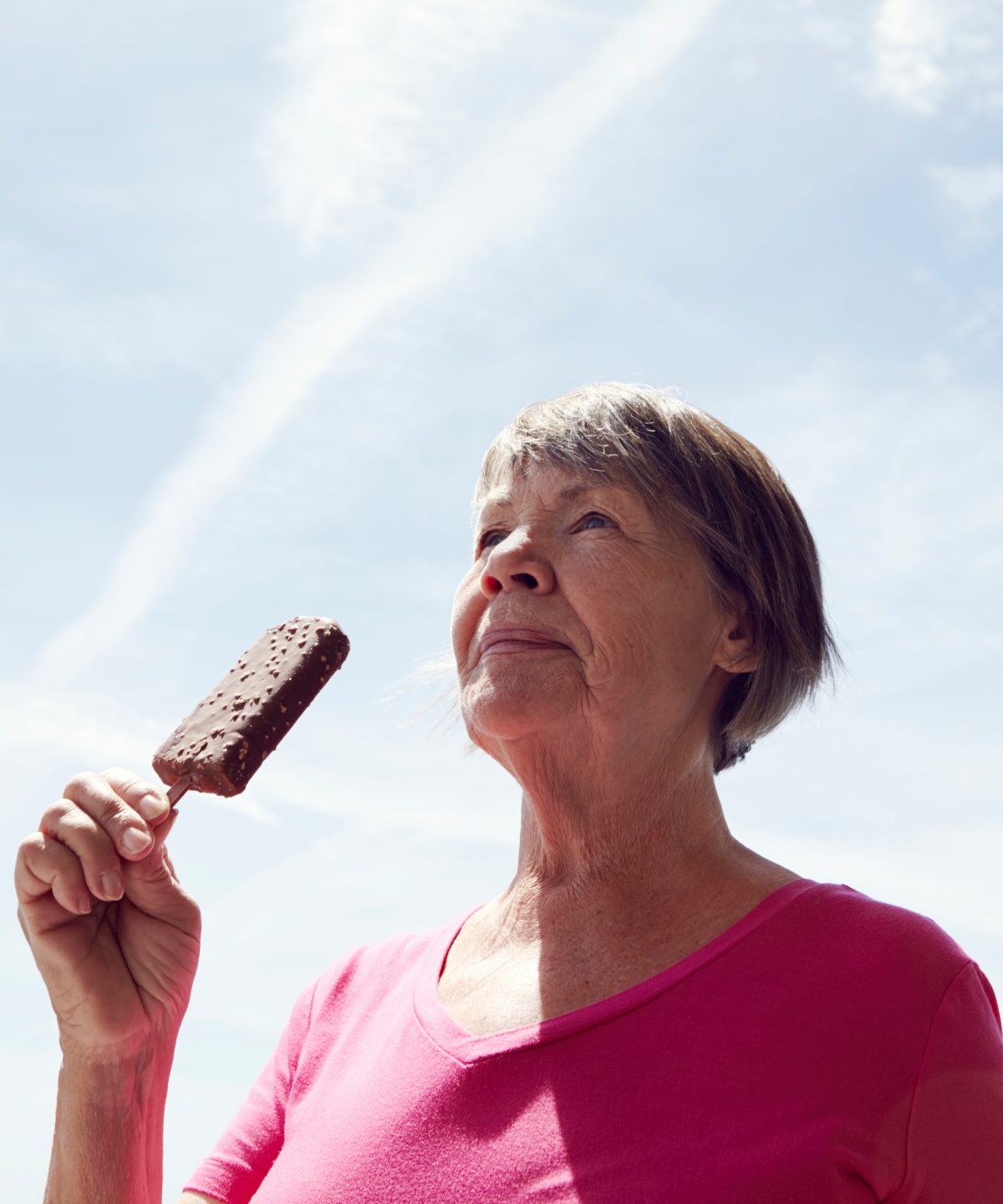 Eine ältere Frau genießt ein Schokoladeneis am Stiel unter blauem Himmel.