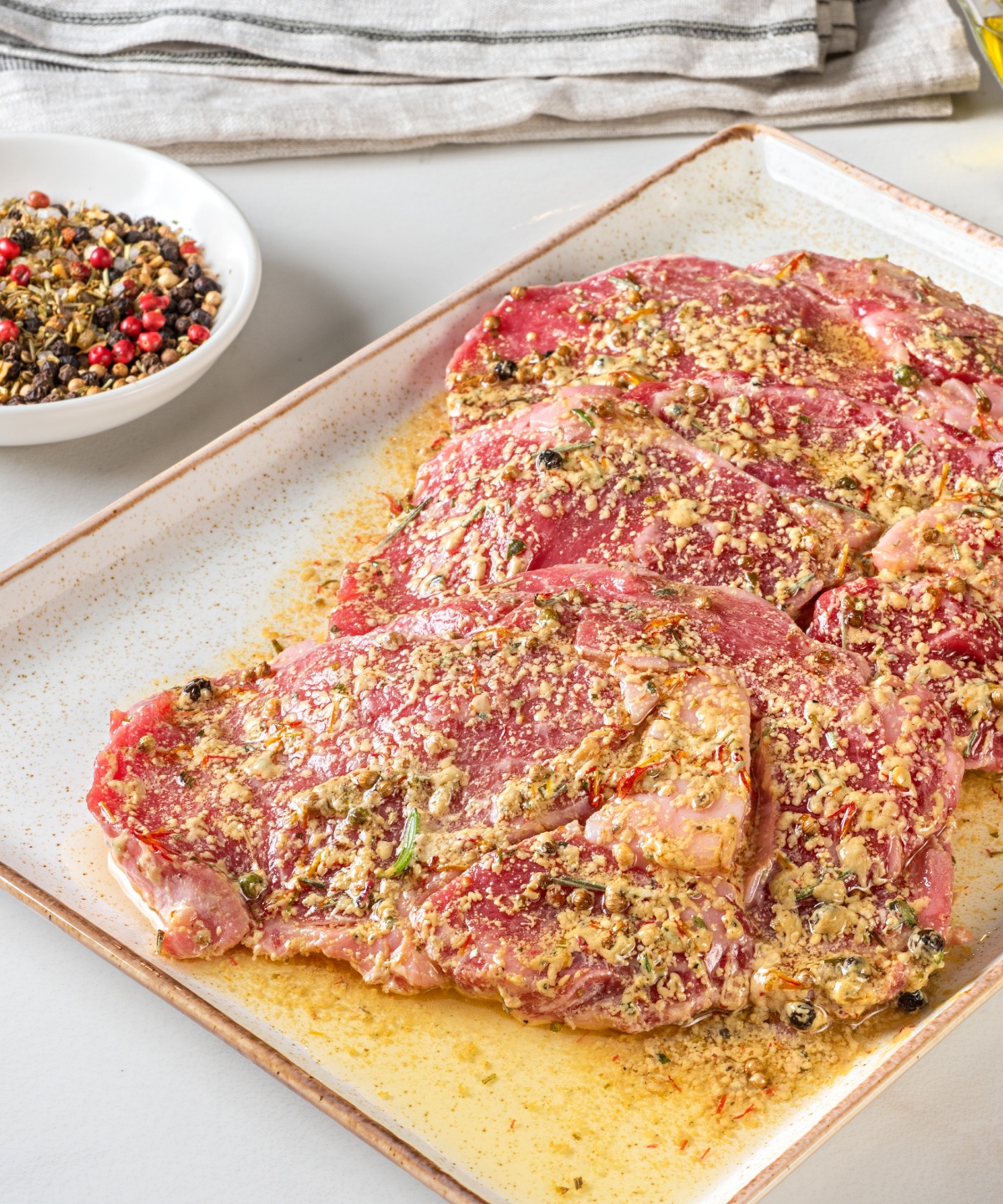 Marinierte Schweinesteaks auf einer Platte, daneben die Zutaten der Marinade wie Öl, Pfeffer, Knoblauch und Rosmarinzweige.