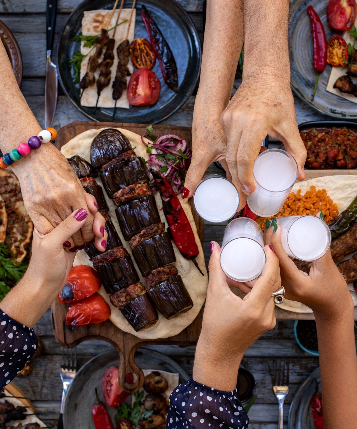 Personen halten sich bei den Händen und stoßen mit Ouzo über einem Tisch mit griechischen Spezialitären an.