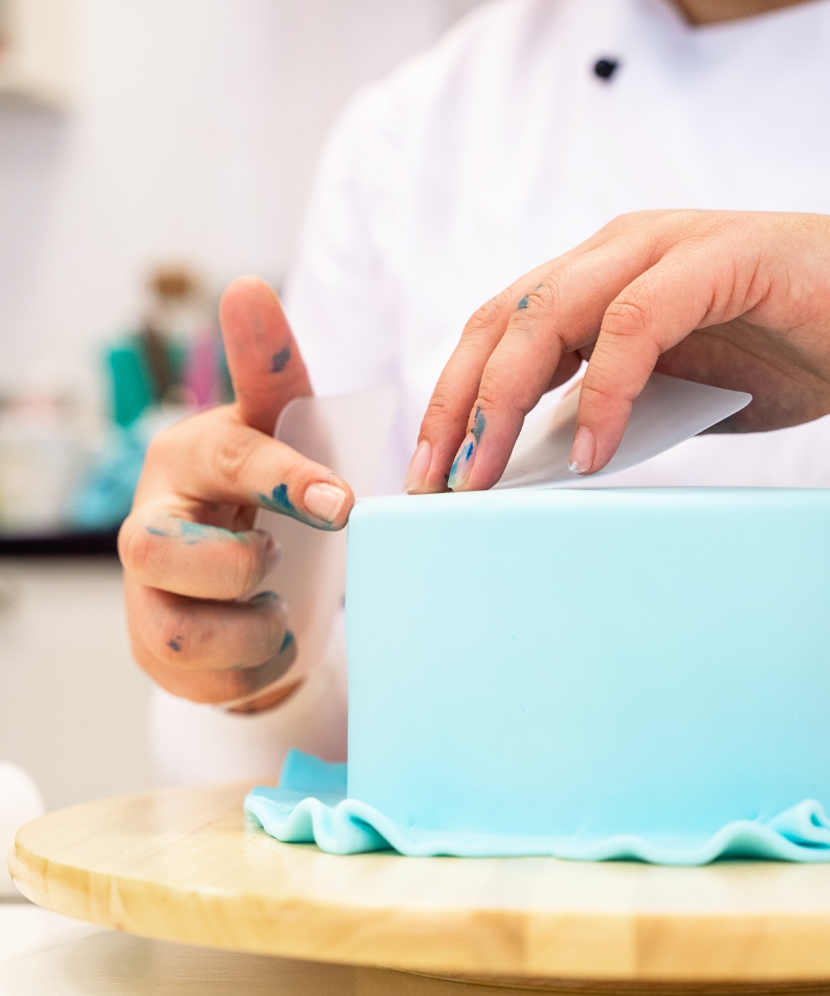 Jemand glättet die Oberfläche einer blauen Fondanttorte mit einem Glättwerkzeug, die auf einem Holzdrehteller steht.