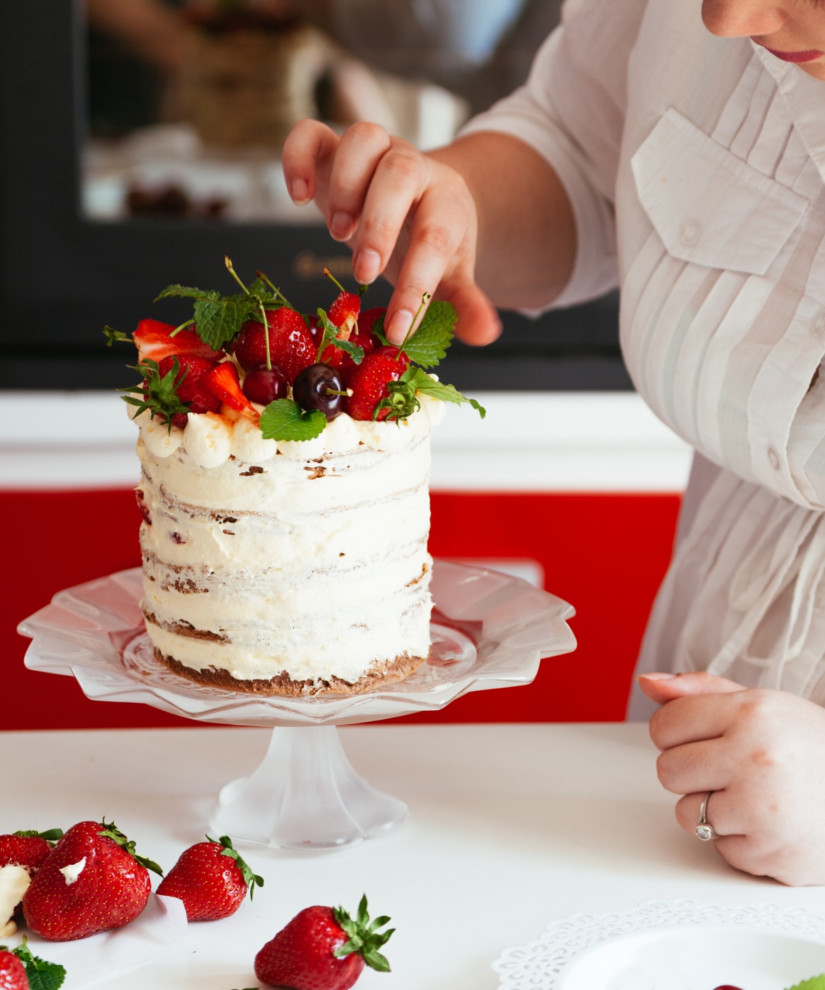 Konditorin dekoriert eine weiße Schichttorte mit Erdbeeren, Kirschen und Minze auf Tortenständer.