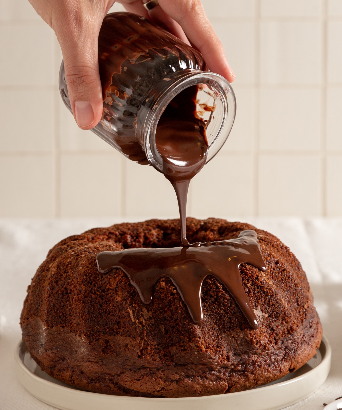 Jemand gießt flüssige Schokolade auf einen Schokoladen-Gugelhupf der auf einem Teller steht.