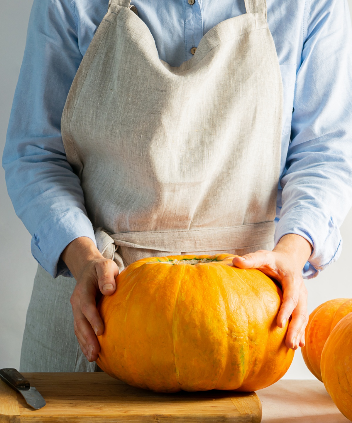 Person in Schürze hält einen großen, runden Kürbis auf einem Schneidebrett. Weitere Kürbisse und Messer daneben.