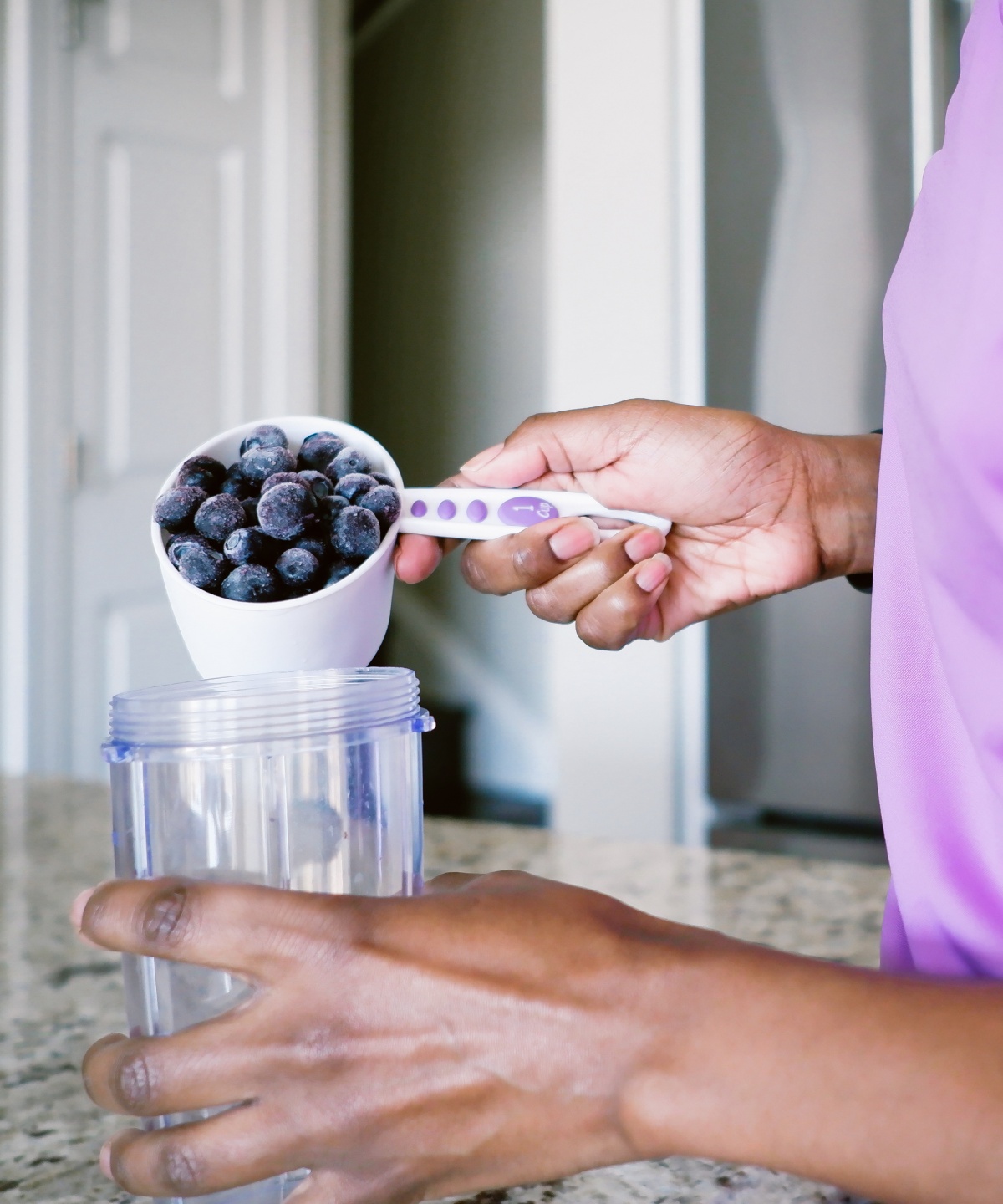Person in lila Shirt füllt gefrorene Blaubeeren aus einem Messbecher in einen Mixbecher.