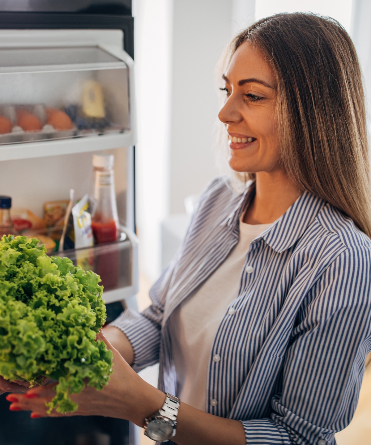 Lächelnde Frau steht vor einem geöffneten Kühlschrank und hält frischen grünen Salat in der Hand.