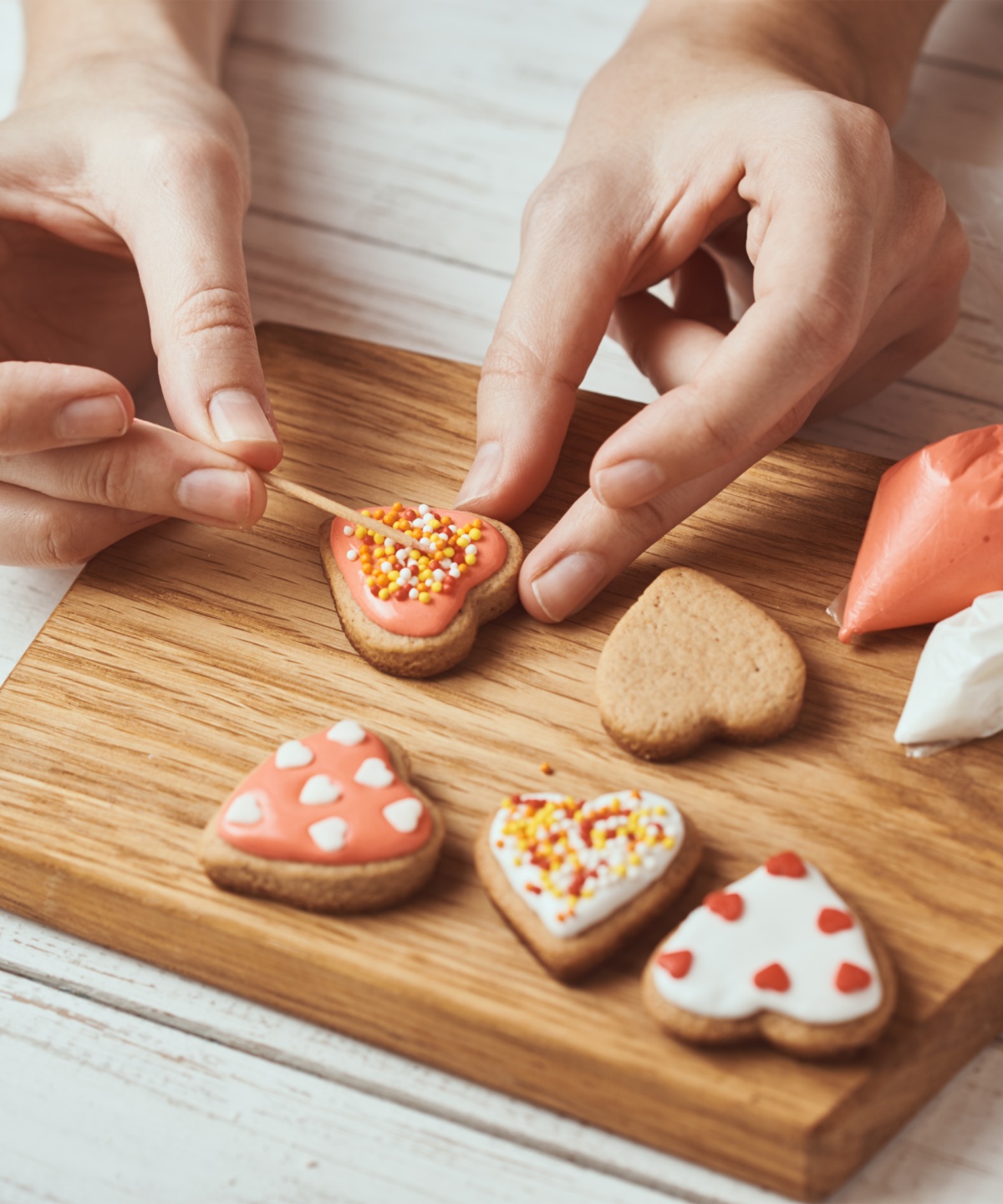 Jemand verziert gebackene herzförmige Plätzchen mit rosa Zuckerguss und Streuseln auf Holzbrett.