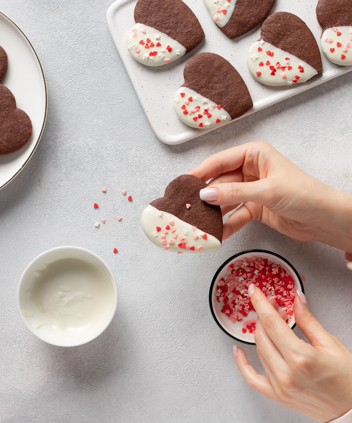 Jemand taucht herzförmiges Schokoladenplätzchen in weiße Glasur und bestreut es mit rosa Streuseln, neben weiteren Plätzchen und Streuseln.