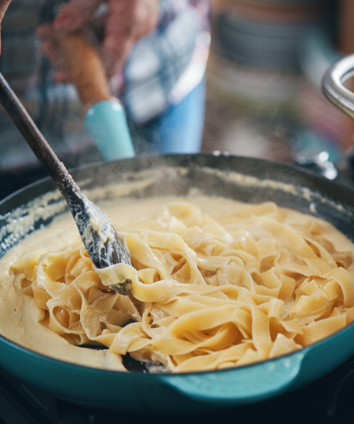 Tagliatelle in cremiger Sauce in einer Pfanne, daneben ein dampfender Kochtopf auf dem Herd.