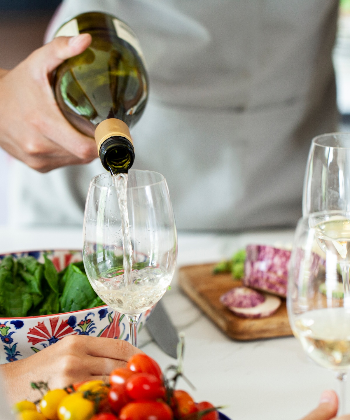 Person gießt Weißwein in ein Glas, während eine Schüssel Salat und geschnittenes Gemüse auf einem Tisch stehen.