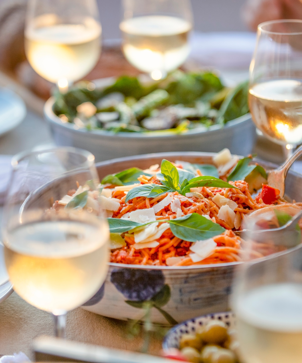 Tisch gedeckt mit Spaghetti in Tomatensoße, Basilikum und Käse, Weißweingläsern und Salat in Schüssel im Hintergrund.