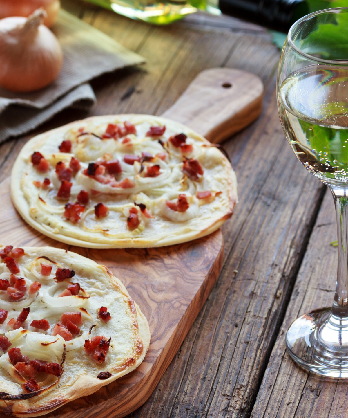 Zwei runde Elsässer Flammenkuchen auf einem Olivenholzbrett, daneben ein Glas Mosel Wein