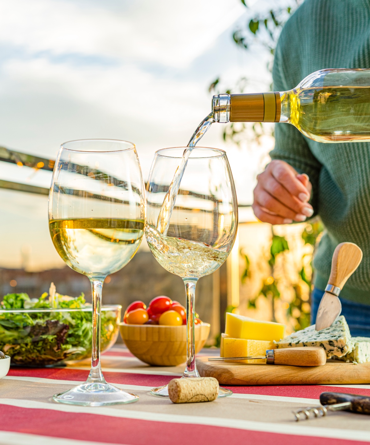 Person schenkt Weißwein in zwei Gläser auf einem Tisch mit Käse, Trauben und Salat bei Sonnenuntergang ein.