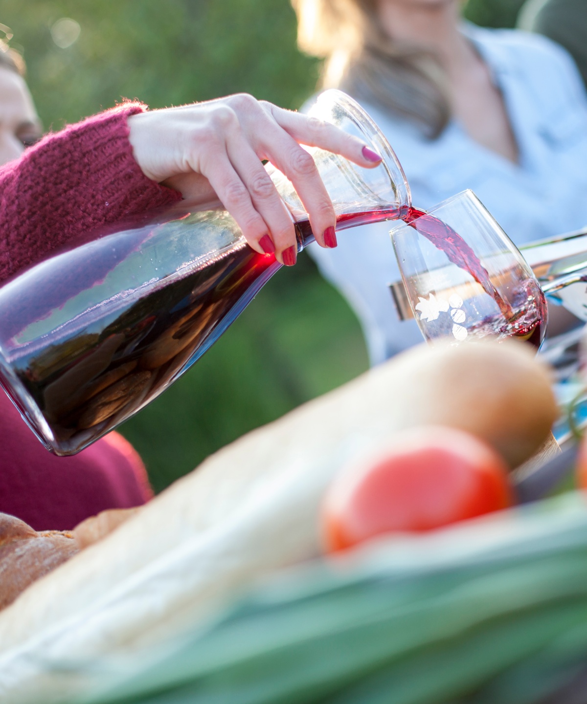 Bei einem geselligen Essen im Freien wird Rotwein aus einer Karaffe in ein Glas gegossen. Im Vordergrund frische Zutaten und Brot.