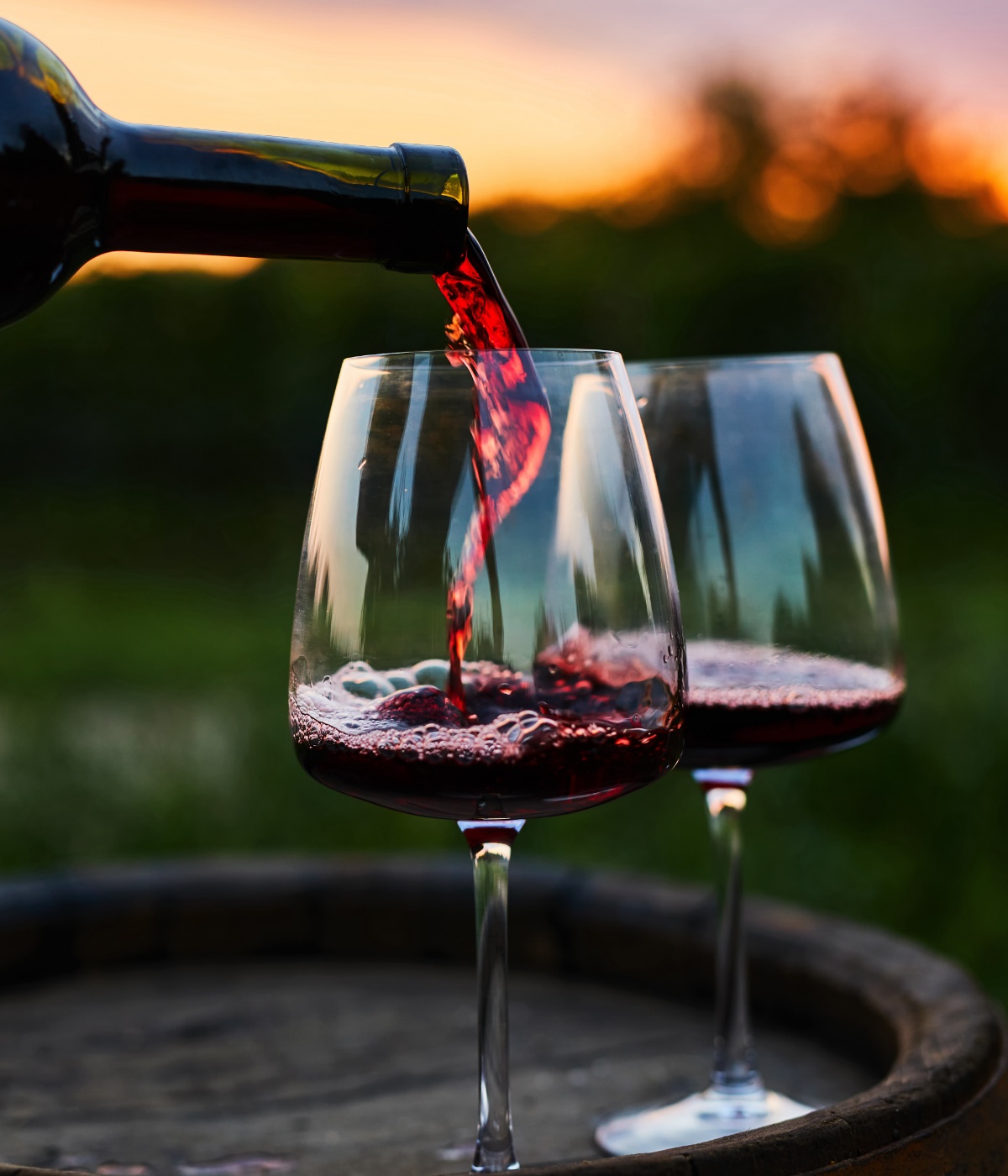 Zwei Weingläser auf einem hölzernen Weinfass werden gerade mit Rotwein gefüllt, im Hintergrund unscharf Natur und Sonnenuntergang.
