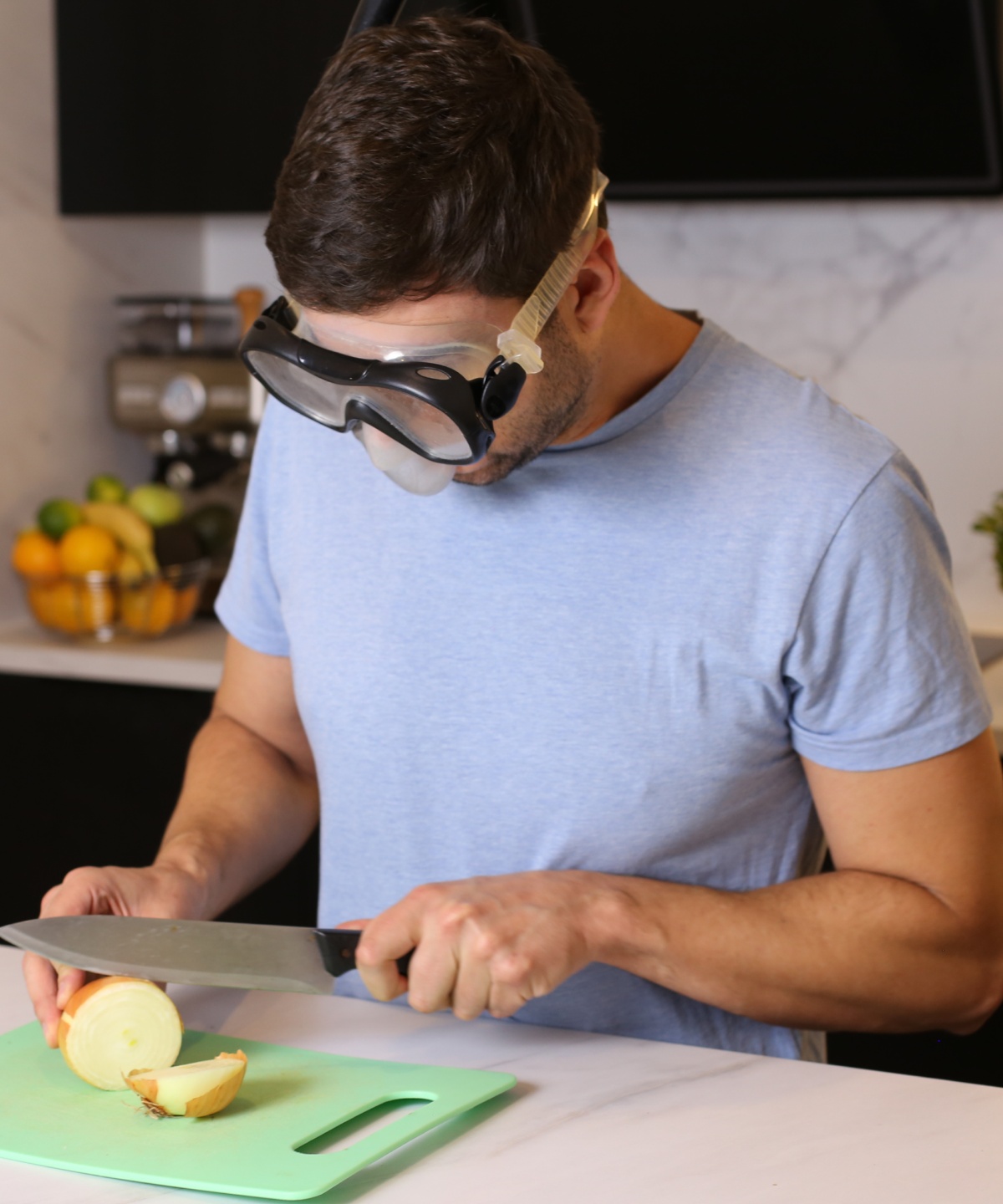 Mann trägt Schutzbrille, während er eine Zwiebel auf einem Schneidebrett in einer modernen Küche schneidet.