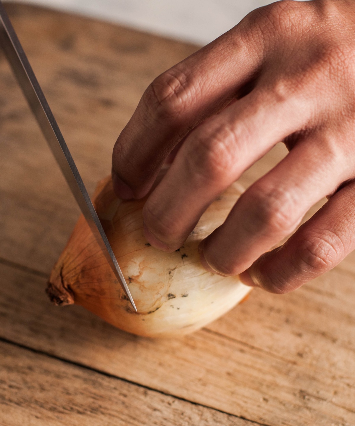Person schneidet eine ungeschälte Zwiebel auf einem rustikalen Holzbrett mit einem scharfen Messer.