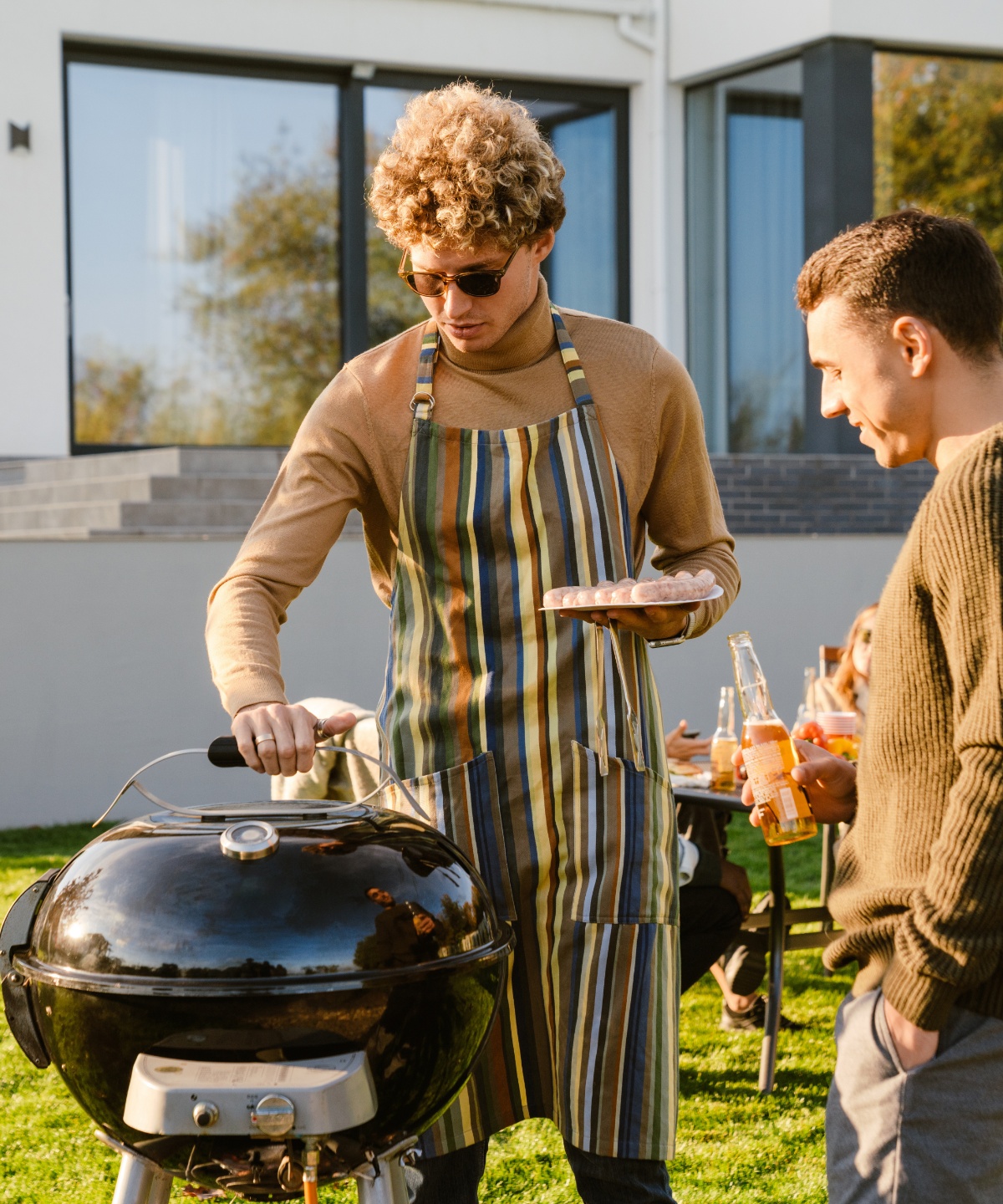 Zwei Männer stehen draußen am Grill, einer mit Sonnenbrille, Schürze und einem Teller mit ungegrillten Würstchen in der Hand, daneben einer eine Flasche Limonade haltend. Im Hintergrund gedeckter Tisch mit Menschen und ein Haus.