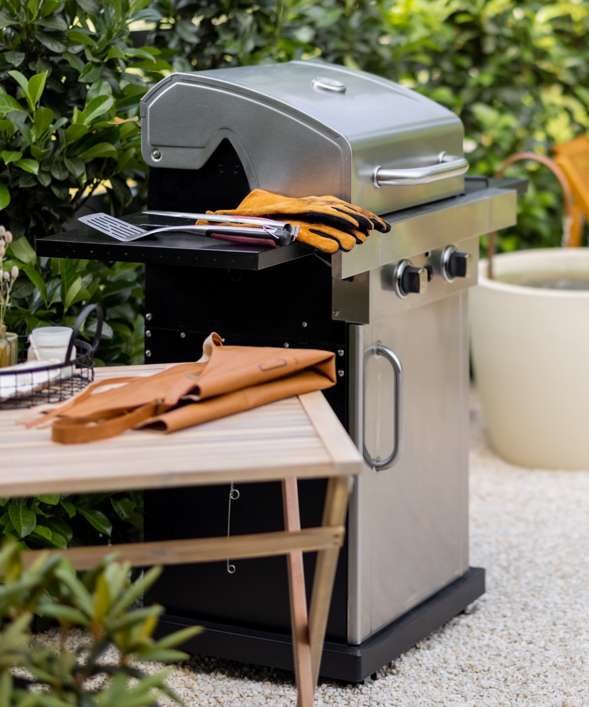Kleiner Holztisch mit gefalteter Schürze darauf, daneben Gasgrill mit Grillhanzschuhen und Grillbesteck, dahinter Heckensträucher.