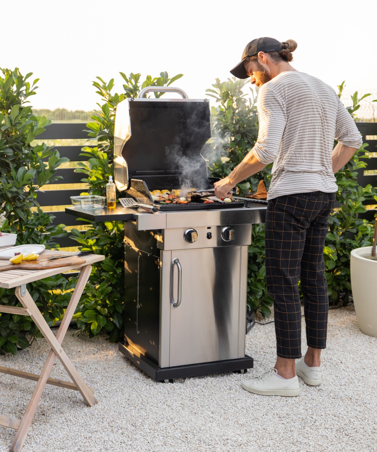 Mann in gestreiftem Shirt, karierter Hose und Schildmütze steht mit Grillzange am mit Gemüse belegten, dampfenden Gasgrill. Auf der Ablage des Grills stehen Ölflasche, leere Aufbewahrungsbox und Pfannenwender. Links davon ein kleiner Holz-Gartentisch mit Schneidebrett und Messer, dahinter Gartenzaun mit Heckensträuchern, dahinter Feld und Bäume.