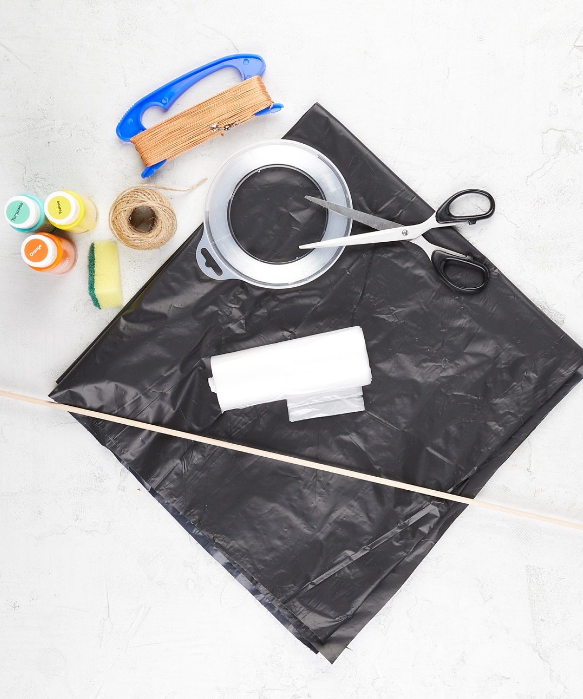 Materialien zum Drachen basteln. Schwarze Müllsäcke, Schere, Klebeband, Schnur, Holzstäbe, Farben und Drachenleine auf hellem Hintergrund.
