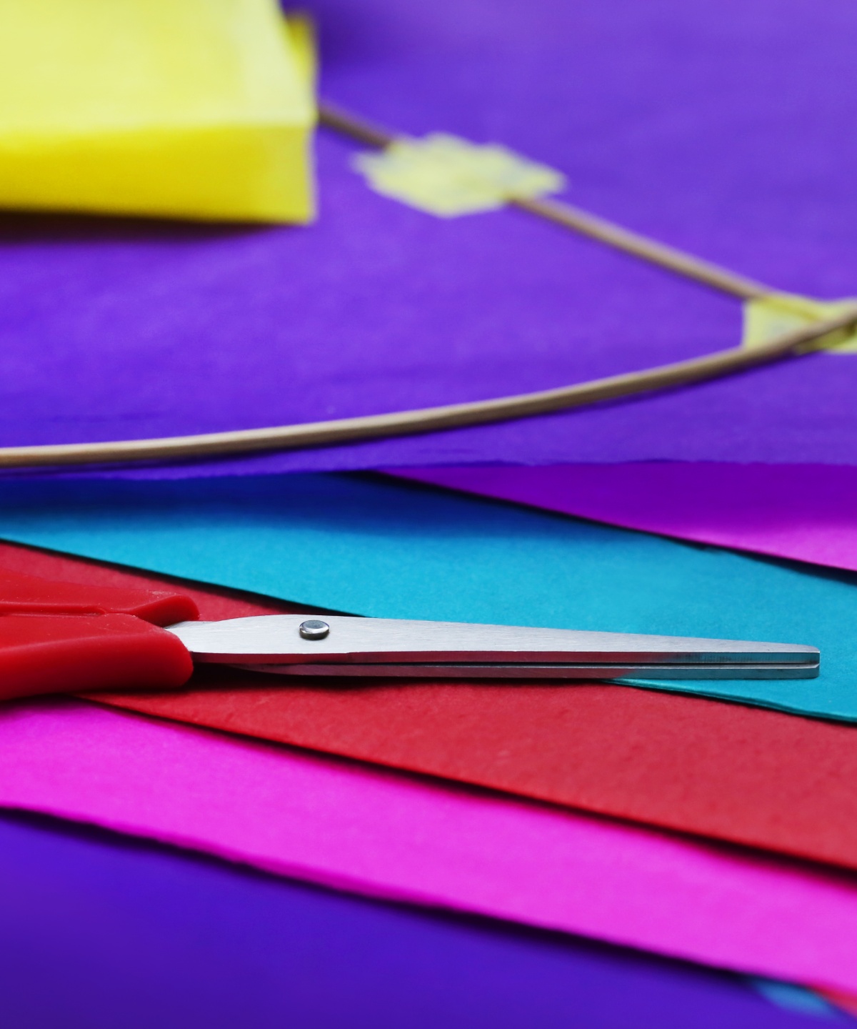 Rote Schere liegt auf buntem Papier neben einem teilmontierten Drachen aus Holzstäben, bereit zum Basteln.