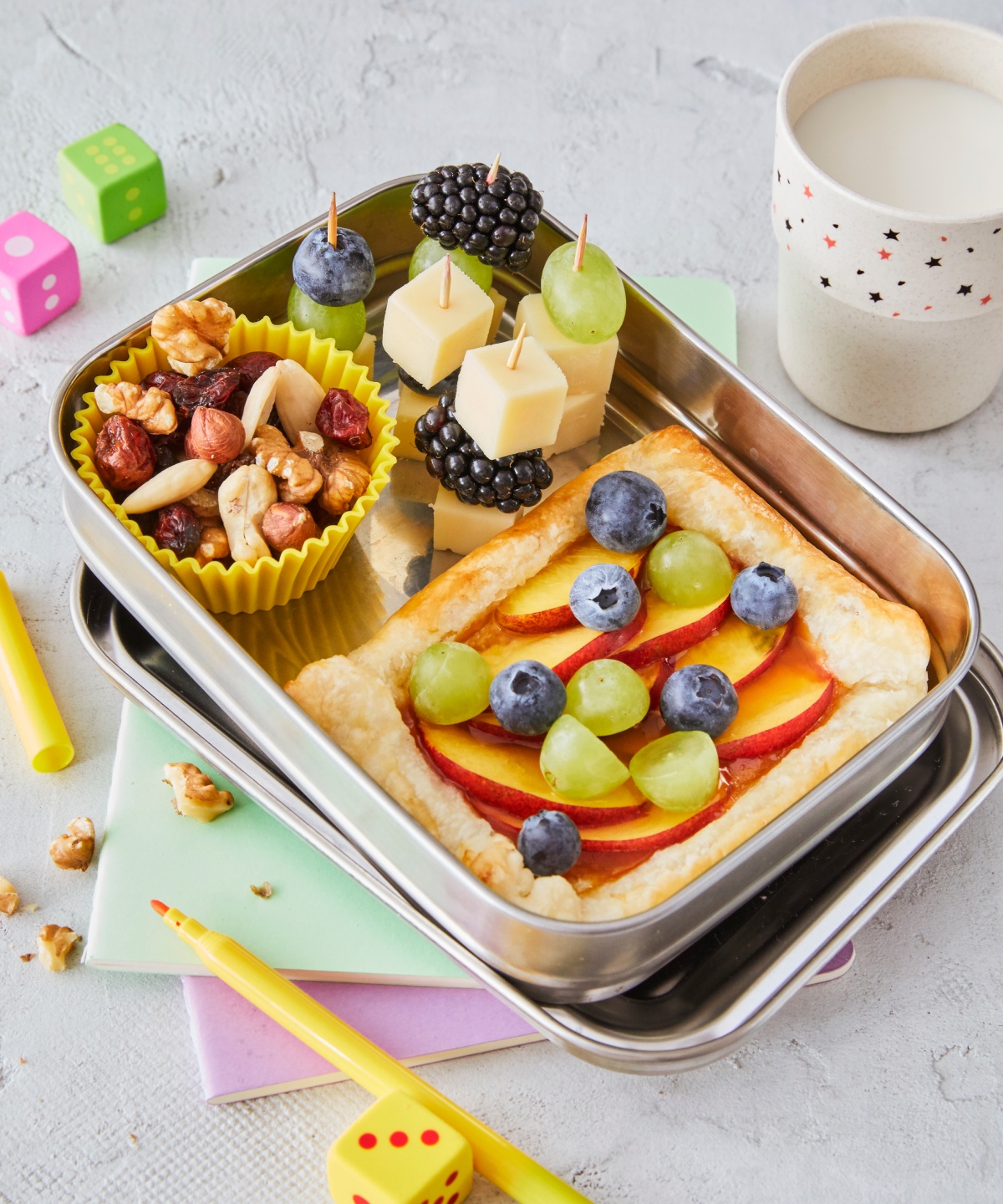 Eine Aluminium-Brotdose, gefüllt mit kleinen Käse-Beeren-Spießen, Nüssen und einer gesunden Obst-Pizza.