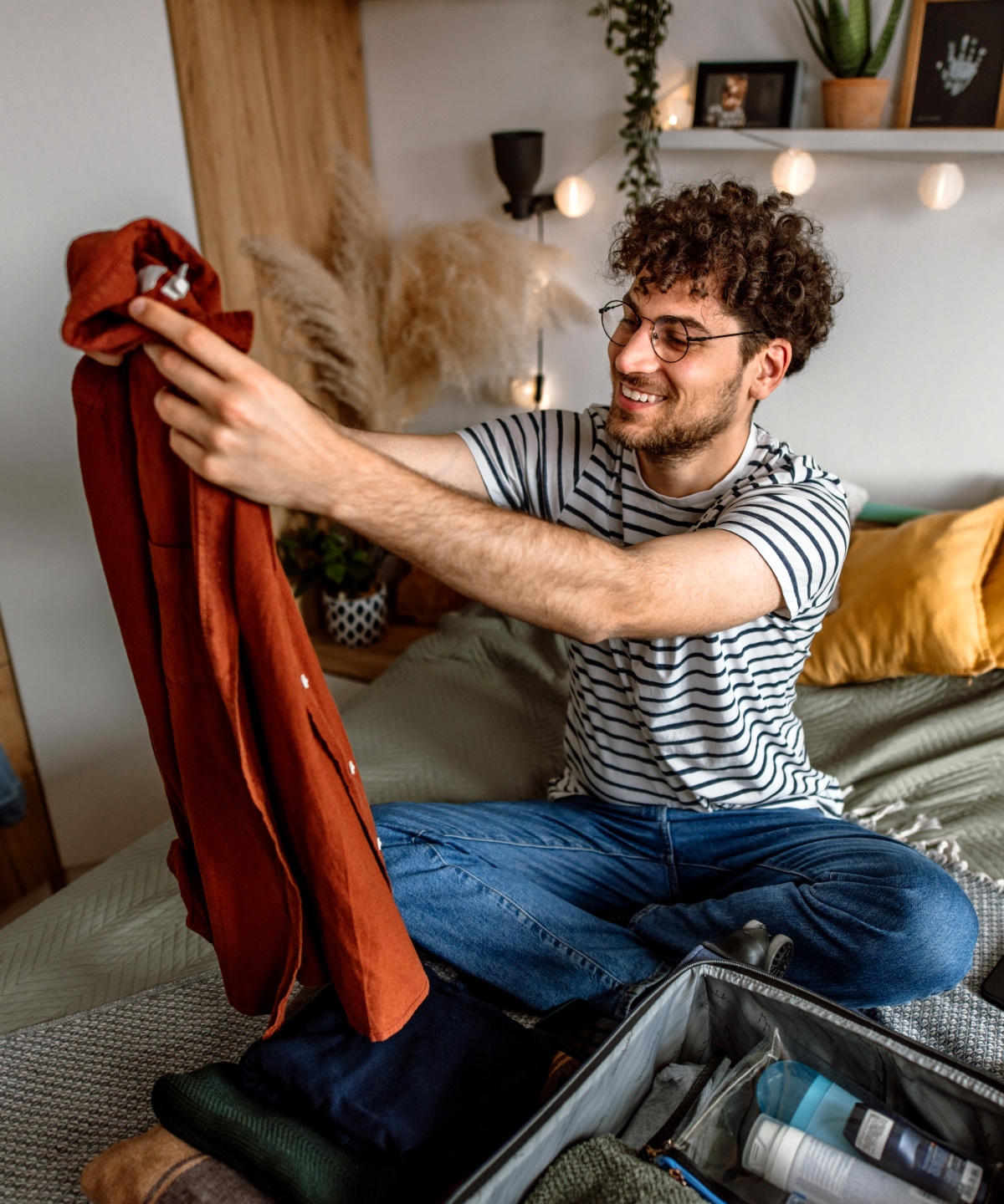 Mann sitzt auf einem Bett beim Kofferpacken. Er hält ein rotes Hemd hoch.
