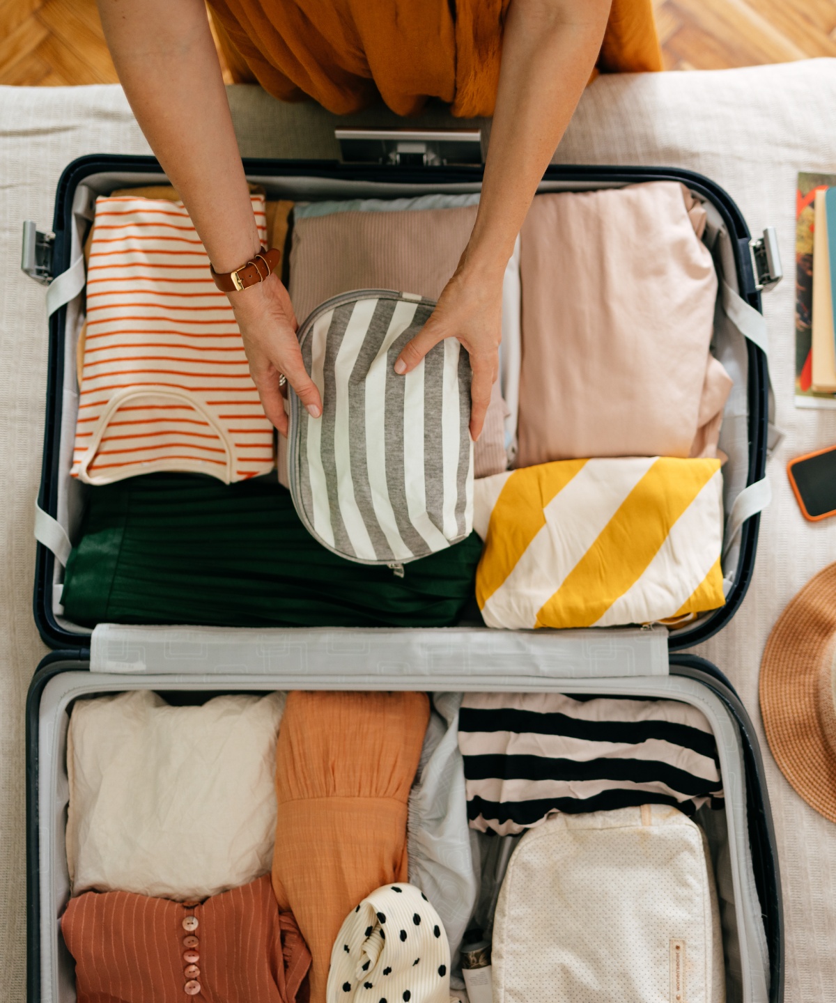 Person packt einen Koffer mit gestreiften Kleidungsstücken, daneben ein Hut, Bücher und andere Dinge.