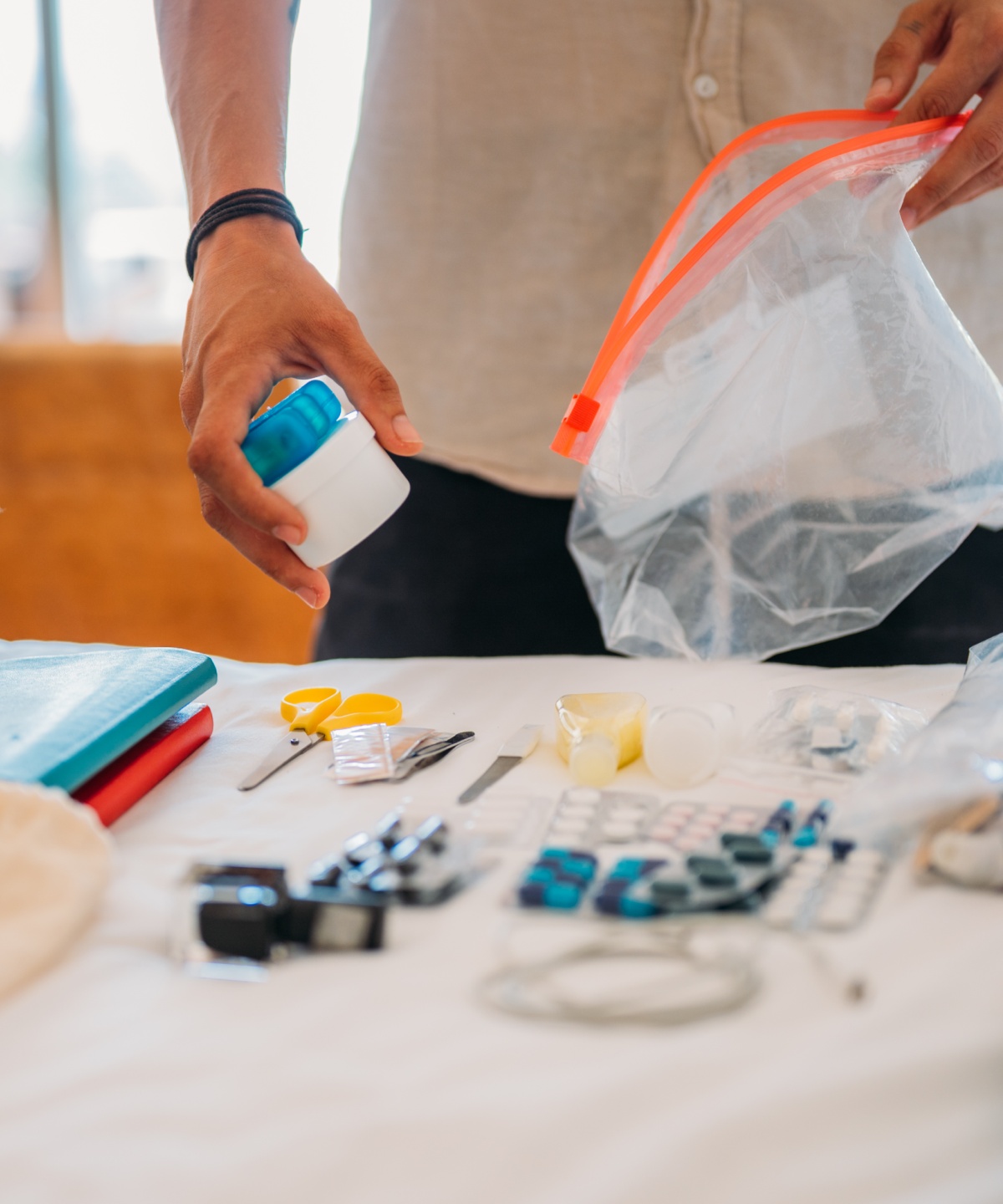 Person packt kleine Gegenstände in einen durchsichtigen Beutel, daneben liegen Kleidung und Bücher.