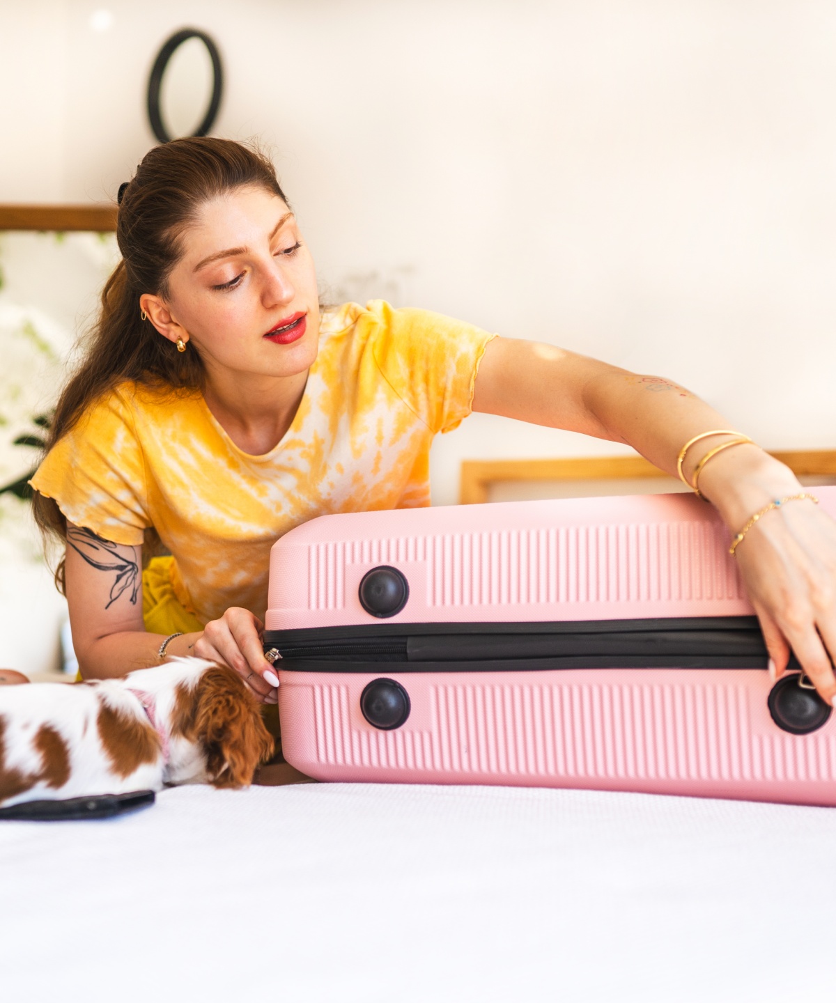 Frau schließt ihren gepackten Koffer auf einem Bett, während ein kleiner Hund daneben liegt.