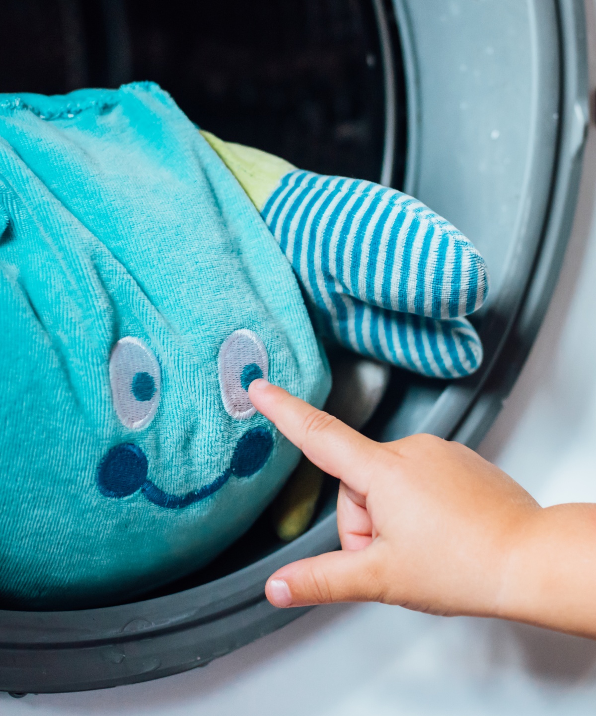 Ein Kuscheltier schaut aus der geöffneten Waschmaschine, während ein Kind ihm einen Stubser gibt.