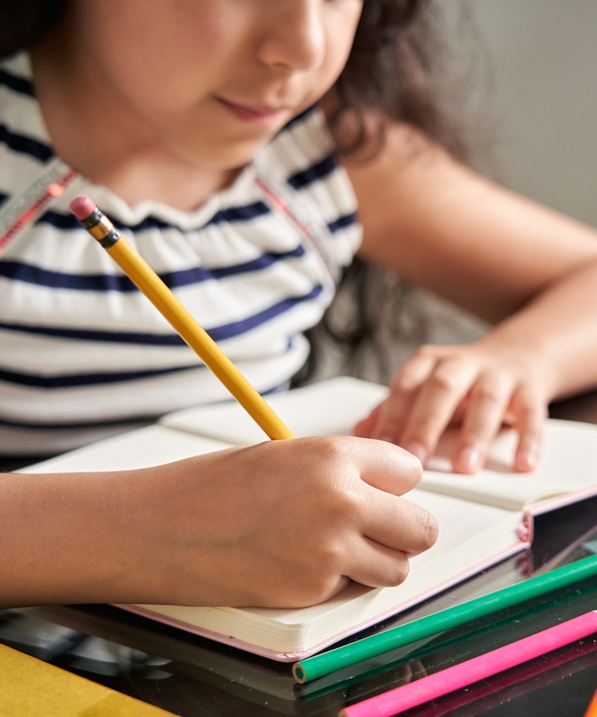Mädchen in gestreiftem Shirt schreibt in ein Notizbuch, auf dem Tisch liegen Buntstifte und weitere Schreibutensilien.