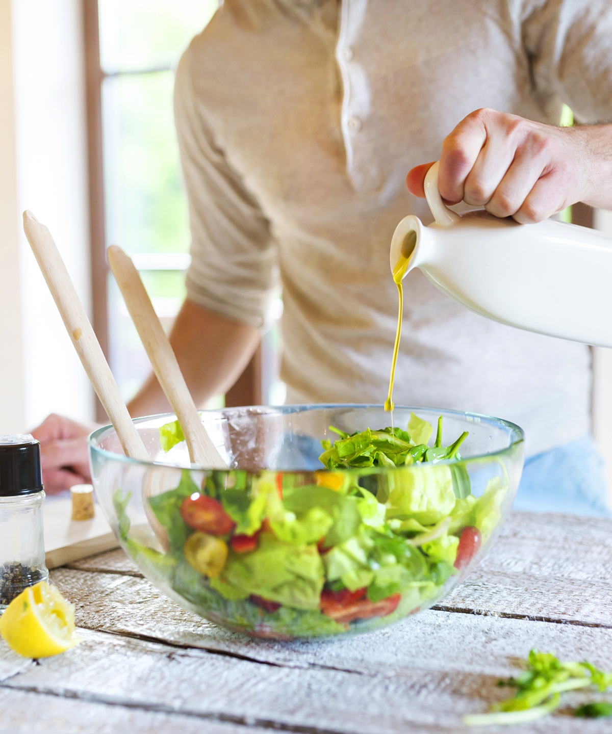 Person gießt Olivenöl in eine Schüssel mit frischem Salat auf einem Holztisch, Gewürze und Zitrone daneben.