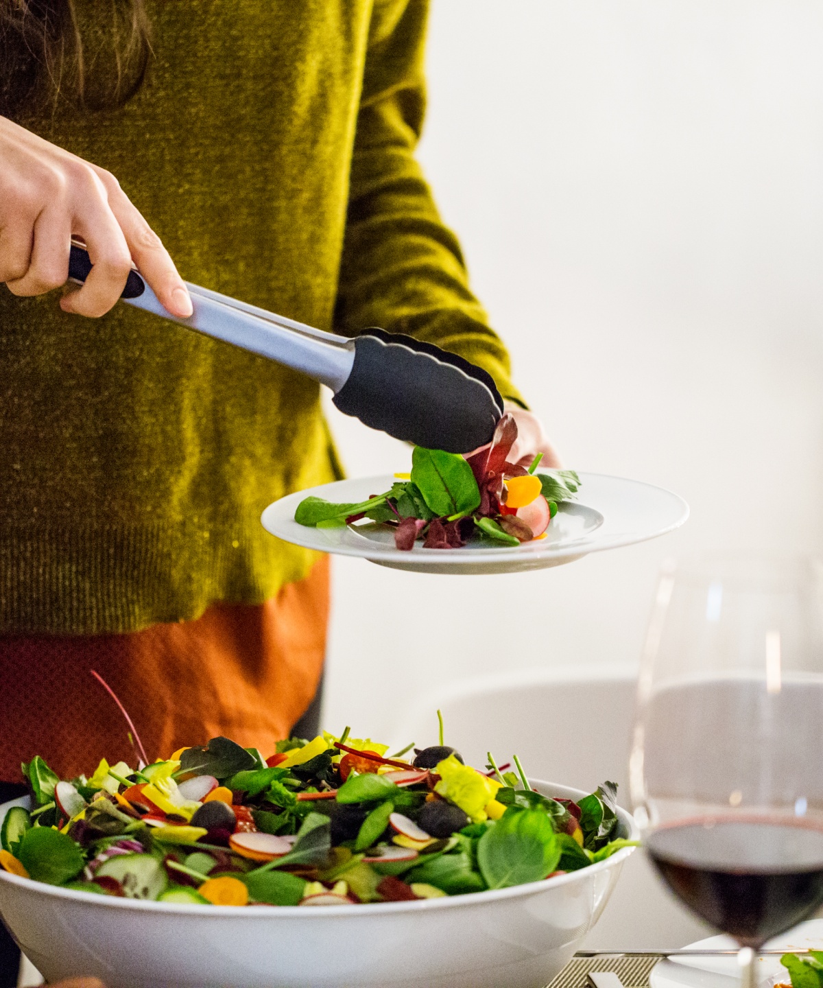 Person serviert frischen Salat auf einen Teller bei einem Abendessen, daneben steht ein Glas Rotwein.