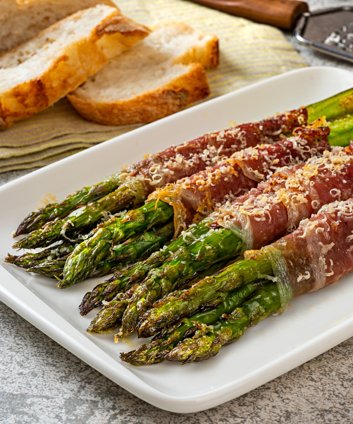 In Speck gewickelter grüner Grillspargel auf einem Servierteller, daneben Brotscheiben und Parmesan.