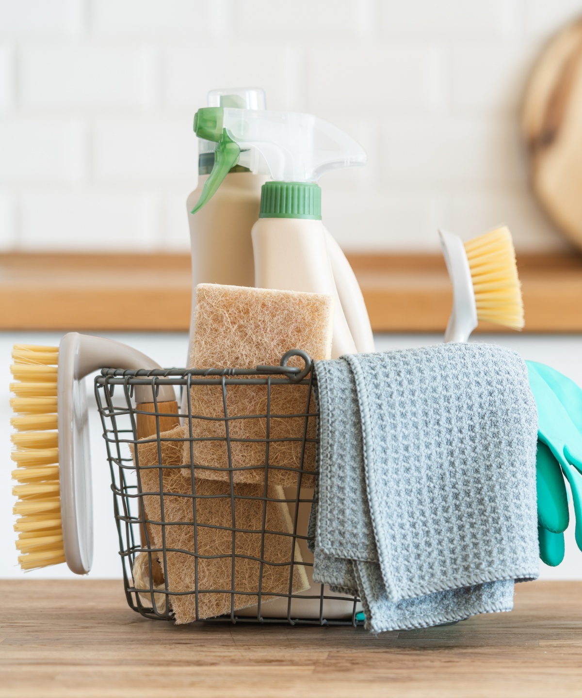 Drahtkorb mit Putzutensilien, wie Schwämmen, Bürsten, Handschuhe und Tücher, die auf einer Holzarbeitsplatte in heller Küche stehen.