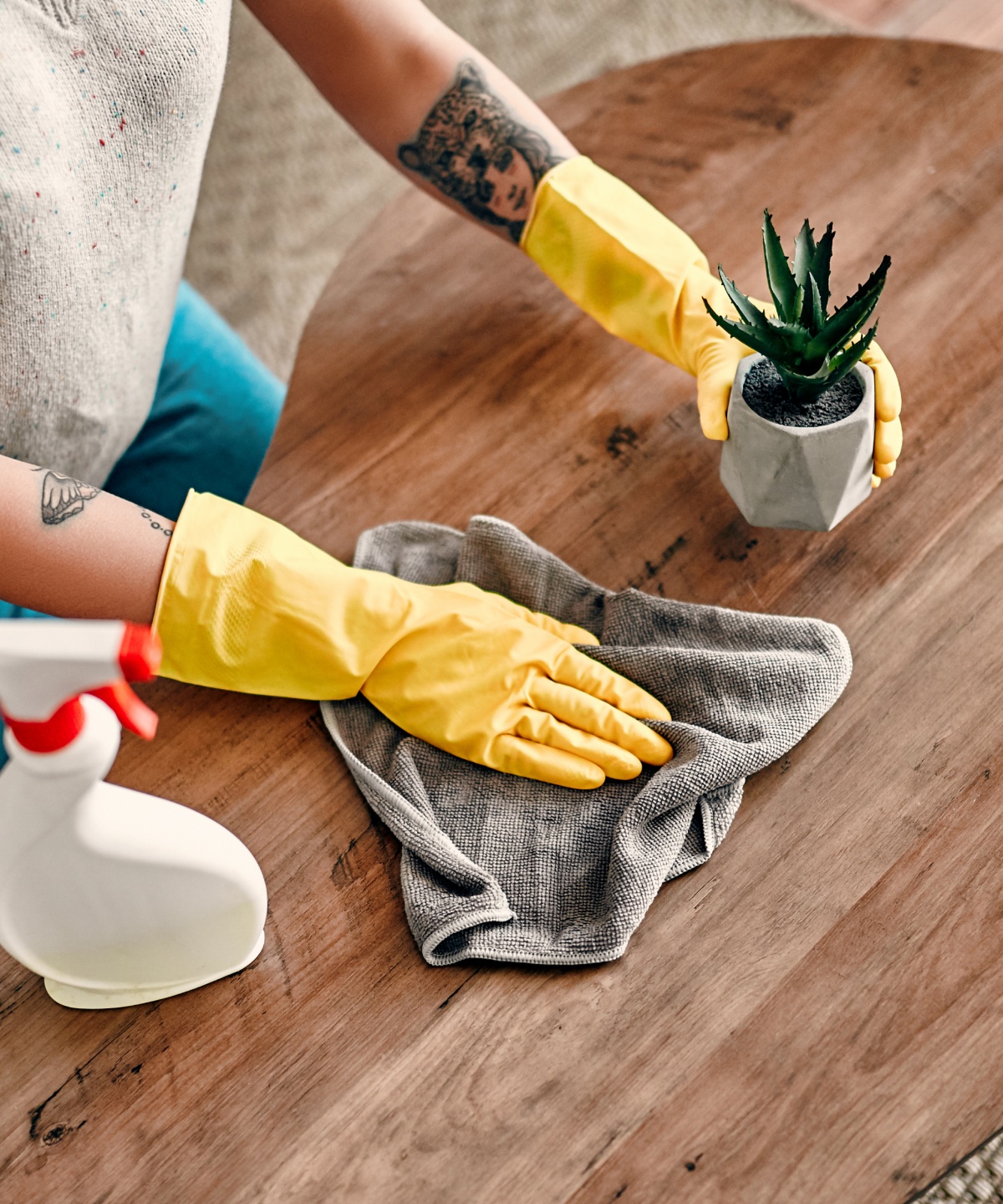 Person mit gelben Handschuhen hebt einen kleinen Blumentopf an, um den Holztisch mit einem Tuch zu reinigen.