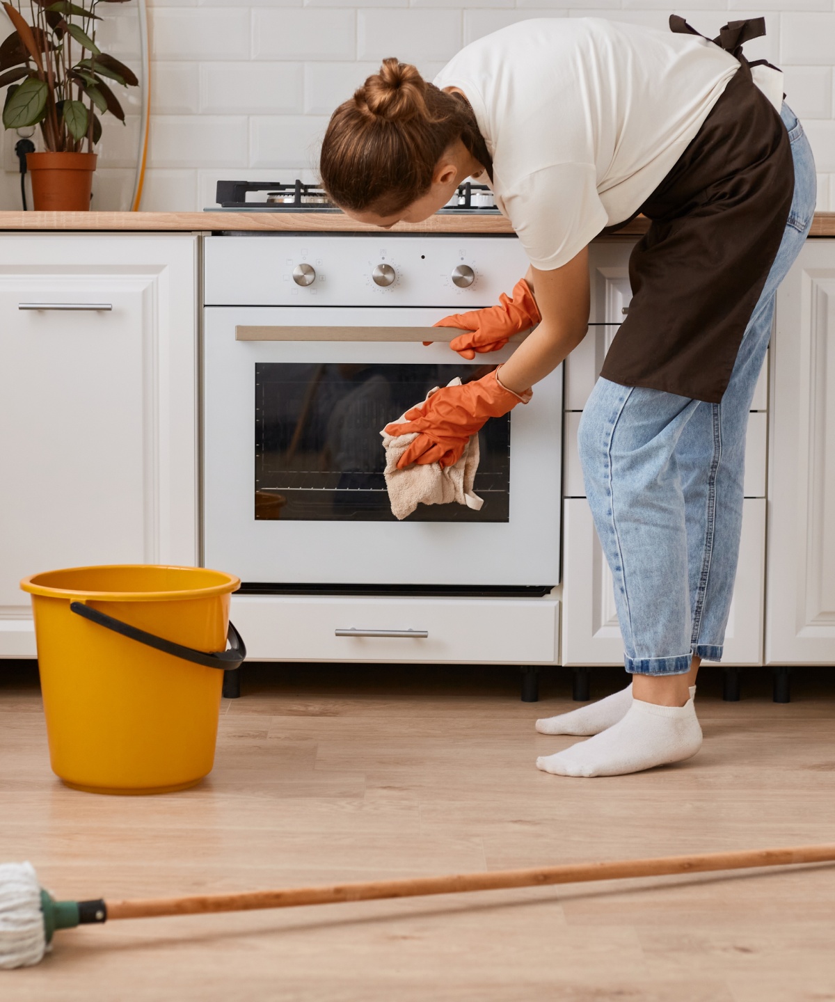 Frau reinigt Ofentür in einer hellen Küche. Auf dem Boden befinden sich Eimer und Wischmopp.