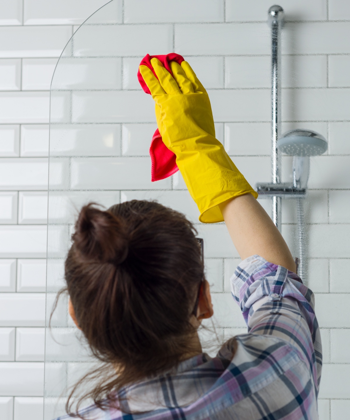 Frau mit gelben Putzhandschuhen reinigt eine Duschwand aus Glas mit einem roten Tuch.