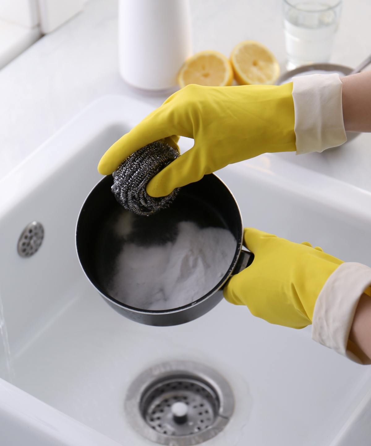 Person mit gelben Putzhandschuhen reinigt Pfanne mit Stahlschwamm in weißem Waschbecken.