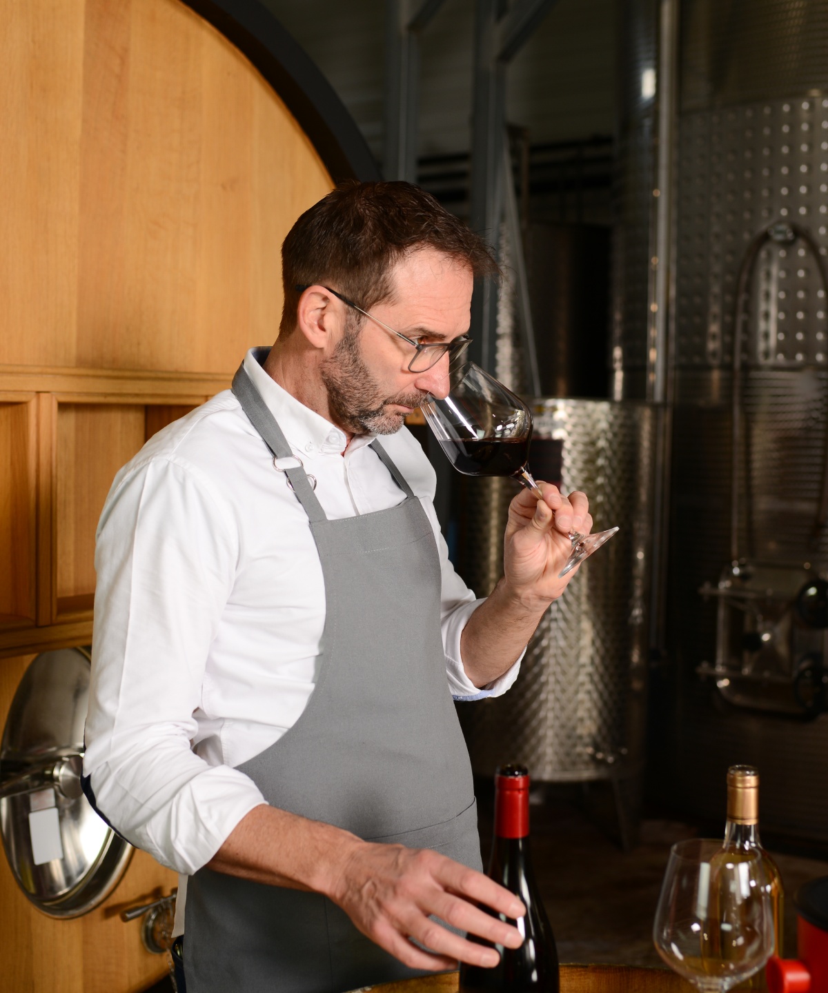 Ein Winzer in Schürze riecht in ein Glas Rotwein. Hinter ihm ein Holzfass und Edelstahlbehälter in einer Weinkellerei.