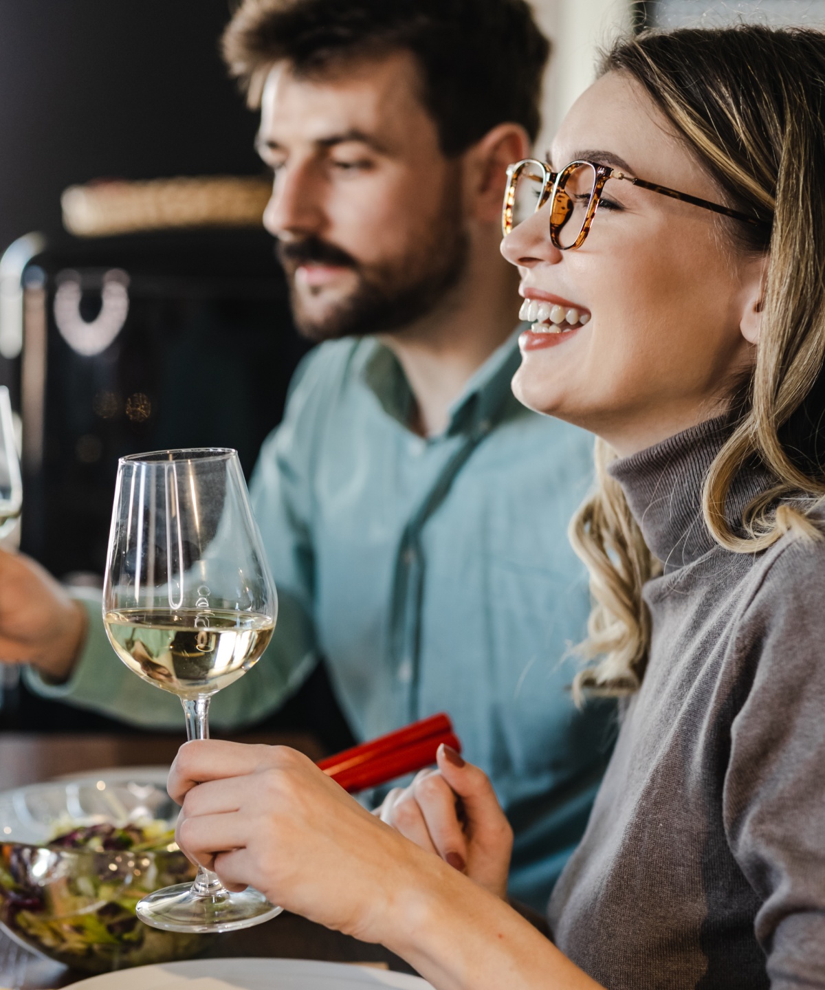 Frau und Mann beim Dinner halten Weißweingläser in den Händen. Auf dem Tische stehen Salat, Käse und Brot.