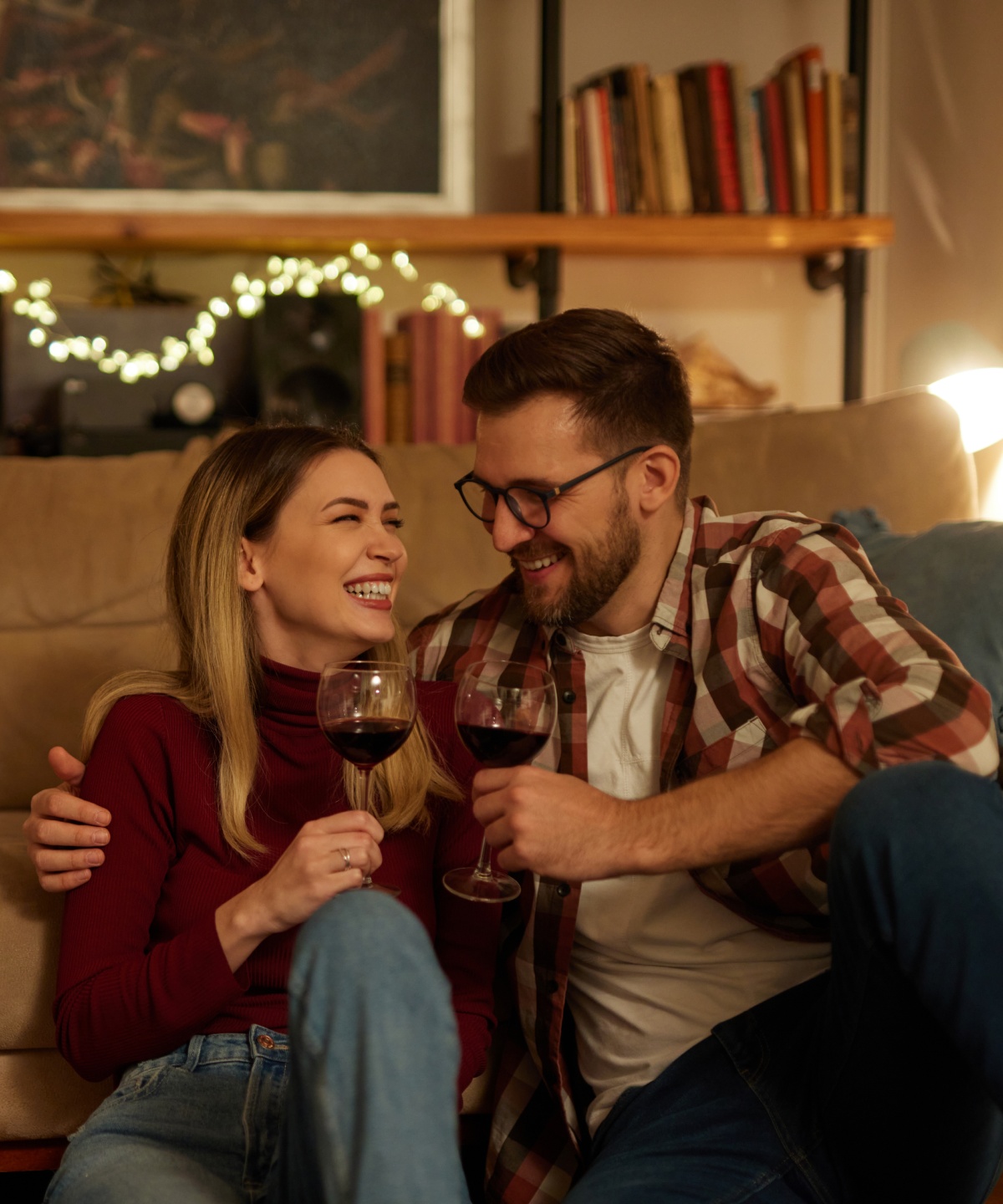 Ein Paar sitzt lachend auf dem Boden vor einem Sofa in einem gemütlichen Wohnzimmer mit gedimmtem Licht. Sie halten Rotweingläser in ihren Händen.