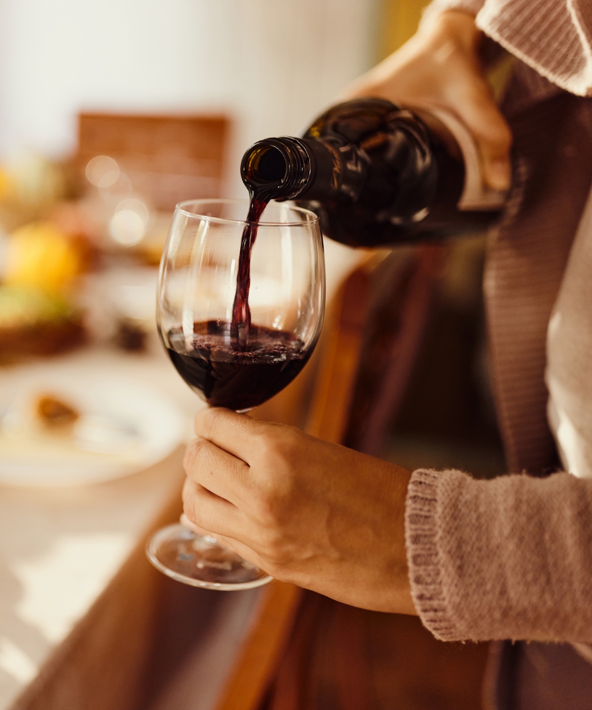 Eine Person schenkt Rotwein in ein Glas ein, im Hintergrund ein gedeckter Esstisch mit verschiedenen Speisen.