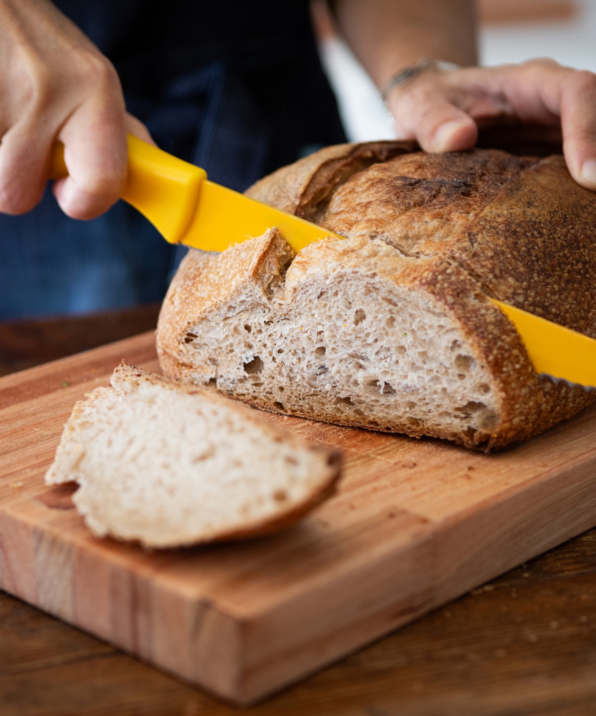 Eine Person schneidet mit einem gelben Brotmesser frisch gebackenes Sauerteigbrot auf einem Holzbrett.