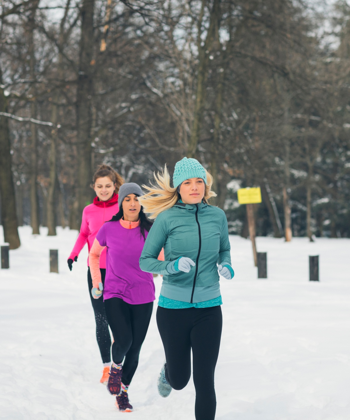 Drei Frauen joggen in bunten Jacken hintereinander durch einen verschneiten Park.