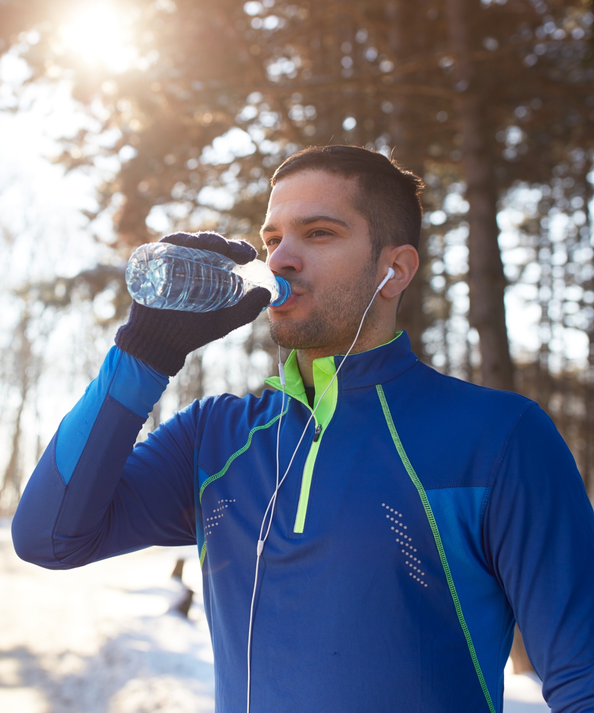 Mann mit blauer Laufjacke und Kopfhörern steht auf einem verschneiten Waldweg und trinkt Wasser aus einer Flasche.
