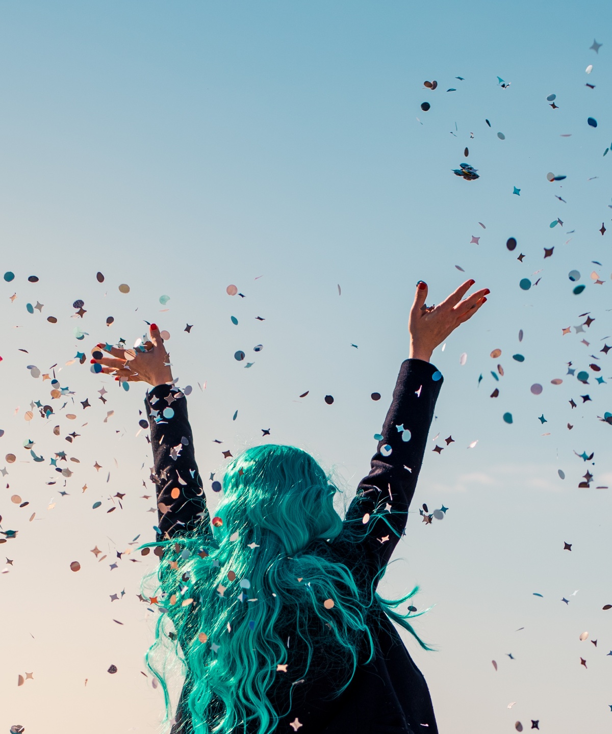 Person mit langen, türkisfarbenen Haaren wirft Konfetti in die Luft bei blauem Himmel im Freien.