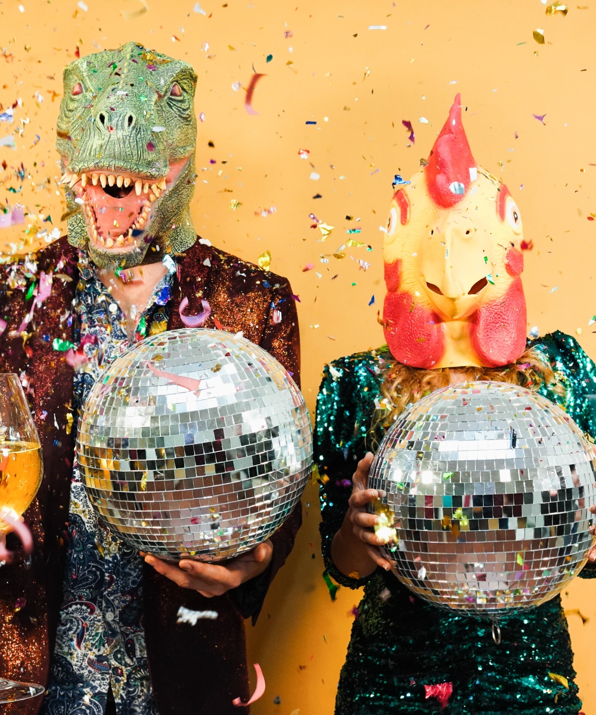 Zwei Personen verkleidet als mit Dino und Huhn, halten große Discokugeln und ein großes Weinglas in der Hand, während Konfetti von oben herunterfällt .